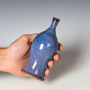 May include: A small ceramic bud vase with a narrow neck and a flared base. The surface features a mottled blue and purple glaze with a glossy finish, while the bottom edge reveals the natural brown clay material of the vessel.
