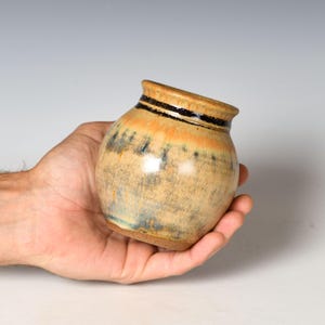 May include: A small, round ceramic pot with a flared rim. The glaze features a blend of warm tan and orange tones, accented by a dark, textured horizontal band near the top and vertical streaks of dark blue and black across the body.