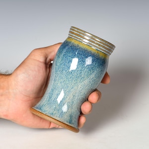 May include: A ceramic tumbler featuring a speckled blue and light blue glaze with a smooth finish. The rim is decorated with horizontal brown and white rings, while the base is unglazed, revealing the natural brown clay texture.
