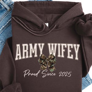 May include: A dark brown hoodie with the text "ARMY WIFEY" in white block letters and a camouflage bow graphic. Below the text is "Proud Since 2025" in a script font. The hoodie is folded on top of blue jeans.
