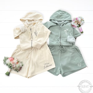 May include: Two matching hooded sweatshirts and shorts sets. One set is cream with the text "Mrs. Johnson" embroidered on the shorts. The other set is sage green with the initials "RJ" embroidered on the shorts.