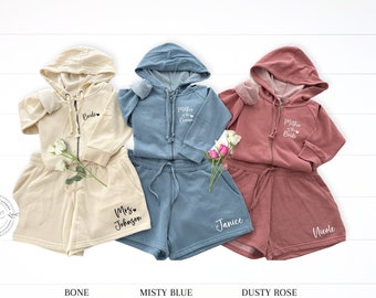 May include: Three sets of zip-up hoodies and shorts in bone, misty blue, and dusty rose colors. Each set features custom white text including Bride, Mrs. Johnson, Mother of the Groom, Janice, Mother of the Bride, and Nicole with small heart icons.