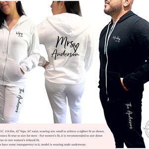 May include: White and black matching zip-up hoodies and joggers. The white hoodie has "Wifey Est. 2024" on the front and "Mrs. Anderson" on the back. The black hoodie has "Hubby Est. 2024" on the front. All joggers have "The Andersons" printed on the leg.