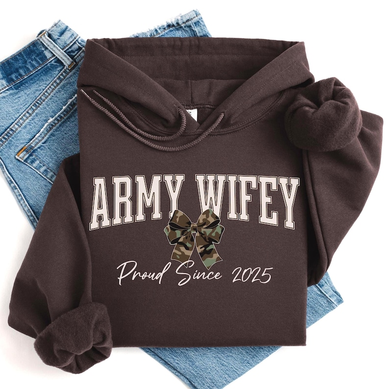 May include: A dark brown hoodie with the text "ARMY WIFEY" in white block letters and a camouflage bow graphic. Below the text is "Proud Since 2025" in a script font. The hoodie is folded on top of blue jeans.