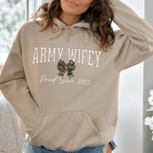 May include: A beige hoodie with a front pocket and drawstring hood. The front of the hoodie features the text "ARMY WIFEY" in a collegiate font, a camouflage bow graphic, and the text "Proud Since 2025".