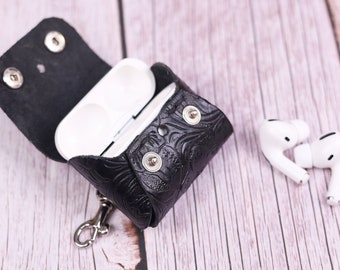 May include: A black leather case with a floral pattern and a silver keychain for carrying wireless earbuds. The case is open and the white earbuds are sitting outside of the case.