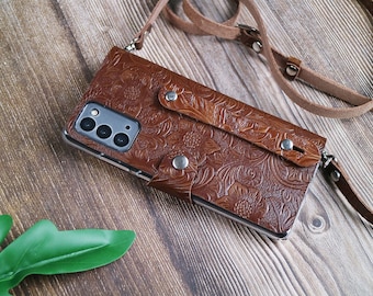 May include: Brown leather phone case with a floral embossed design. The case has a strap and metal snap closures. The phone case is designed to hold a smartphone and has a camera cutout.