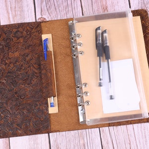Puede incluir: Un cuaderno encuadernado en cuero marrón con un diseño en relieve floral. El cuaderno tiene un bolsillo de plástico transparente en el lado derecho y un portaplumas en el lado izquierdo. Dos bolígrafos negros y un bolígrafo azul son visibles en los bolsillos.