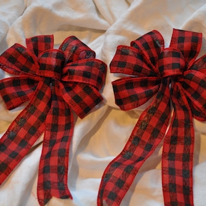 May include: Two decorative bows with a red and black buffalo plaid pattern. Each bow has multiple loops and long, flowing tails. The bows are made of a sheer fabric and are set against a white background.