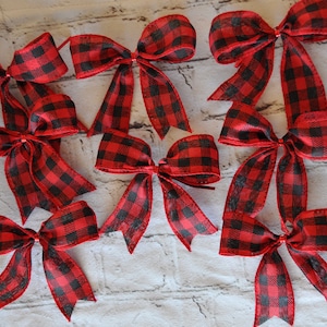 May include: Ten red and black buffalo plaid bows with wired tails. The bows are arranged in a semi-circle on a white fabric background.