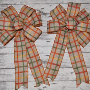 May include: Two decorative plaid bows with long tails. The bows feature a tan background with a plaid pattern in orange, green, and yellow. The edges of the bows are trimmed in orange.