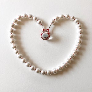 Puede incluir: Un collar de perlas blancas con un colgante de vidrio rojo y blanco en forma de corazón. El collar está dispuesto en forma de corazón sobre un fondo blanco.