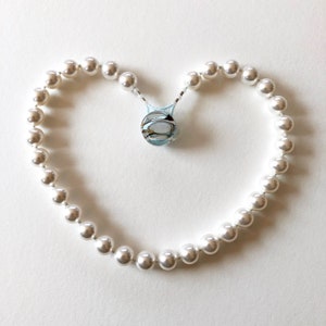 Puede incluir: Un collar de perlas blancas en forma de corazón. El collar tiene un cierre plateado.