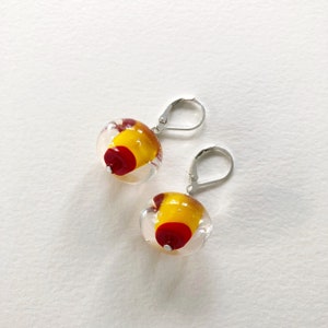 May include: A pair of clear glass earrings with a yellow and red design. The earrings have a lever back closure made of silver-colored metal. The glass orbs are round and have a unique, colorful pattern.
