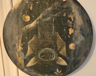 Könnte beinhalten: Ein rundes Hängeschild aus Schiefer mit einer handgemalten Illustration einer Fledermaus, die kopfüber an einem Ast hängt. Das Bild zeigt zwei kleine Kürbislaternen, goldfarbene Akzente und kleine, klare Strasssteine. Es hängt an einer Juteschnur.