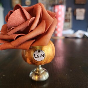 May include: A decorative floral arrangement featuring a large, rust-colored rose atop a gold-toned, pedestal-style base. A small silver charm with the word "Love" hangs from the base. The arrangement sits on a dark wooden table.