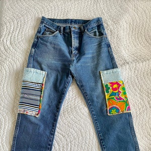 May include: Blue denim jeans featuring rectangular fabric patches sewn onto the front of each leg. The left patch displays horizontal blue, white, and black stripes, while the right patch features a vibrant floral pattern in orange, pink, and green.