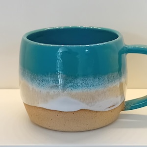 May include: A ceramic mug featuring a glossy teal upper section that transitions into a white band, followed by a textured, unglazed beige base. The mug has a rounded body and a matching teal handle on the right side, creating a distinct colour-blocked appearance.