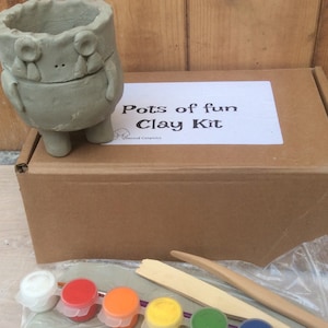 May include: A grey clay kit featuring a frog-shaped pot, a cardboard box with the text "Pots of fun Clay Kit", and painting supplies. The kit includes paint pots in various colours, clay, and wooden sculpting tools.