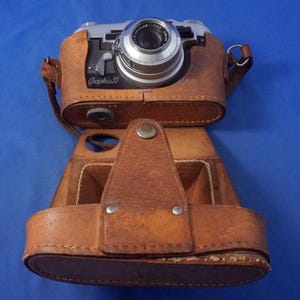 May include: A vintage Graphic 35 camera with a silver lens and black body, encased in a tan leather protective case. The camera features a prominent lens assembly with visible text and a metal shutter release lever on the front panel.