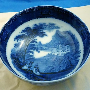 May include: A blue and white ceramic bowl with a landscape scene. The bowl features a detailed illustration of a castle, trees, and figures. The rim of the bowl has a decorative blue pattern.