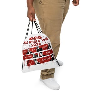 Puede incluir: Bolsa con cordón blanca con el texto "BTS WORLD TOUR 2026" en rojo y negro. La bolsa muestra imágenes de los miembros de la banda y sus nombres. El diseño incluye salpicaduras rojas.