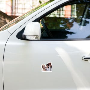 May include: A sticker featuring a group of three people, with a light-skinned woman on the left, a dark-haired person in the middle, and a person with dark hair on the right. The sticker is affixed to the white exterior of a car.