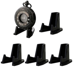 May include: Five black plastic display stands for pocket watches. The stands are shaped like a triangle with a raised platform in the center. The stands are all the same size and shape. One stand has a pocket watch on it. The pocket watch is black with a silver face and Roman numeral markings.