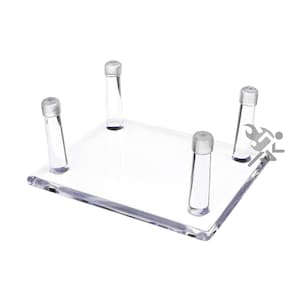 May include: Clear acrylic display stand with four clear plastic feet. The stand is square shaped and has a smooth surface.