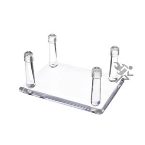 May include: A clear acrylic display stand with four cylindrical supports. The stand is rectangular and designed to showcase small items. The supports are evenly spaced and capped with rounded tops.