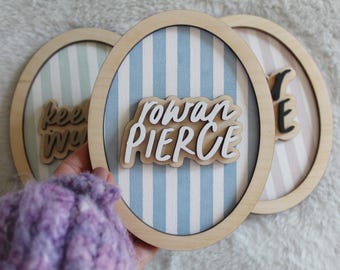 Letrero ovalado de madera con diseño de rayas de acuarela azul y nombre de bebé personalizado.