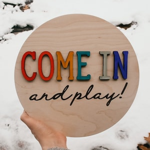 K&ouml;nnte beinhalten: Runder Holzschild mit dem Text "COME IN and play!" in farbigen, erhabenen Buchstaben. Der Hintergrund ist verschneit. Die Buchstaben sind rot, orange, t&uuml;rkis, grau und blau.