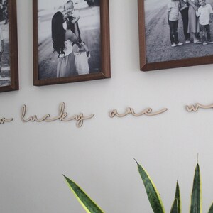May include: Wooden wall decor with the words "how lucky are we" in a cursive font. The decor is displayed on a white wall with framed photos of families.