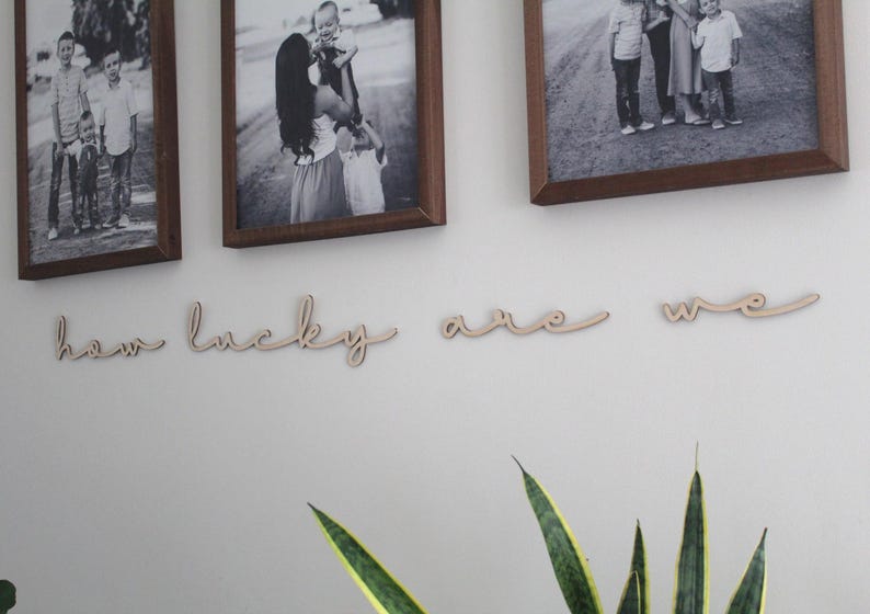 May include: Wooden wall decor with the words "how lucky are we" in a cursive font. The decor is displayed on a white wall with framed photos of families.
