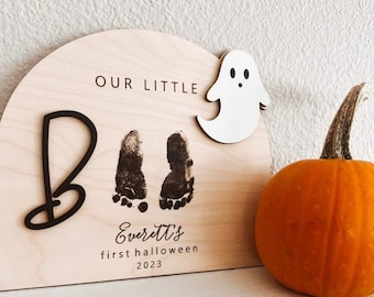 以下が含まれることがあります： 幽霊の切り抜き、赤ちゃんの足跡2つ、そして「Our Little Everett's First Halloween 2023」という文字が書かれた木製の看板です。