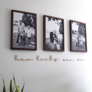 May include: Three black and white framed photos of a family hanging on a white wall with the words "how lucky are we" written in wood below the photos.