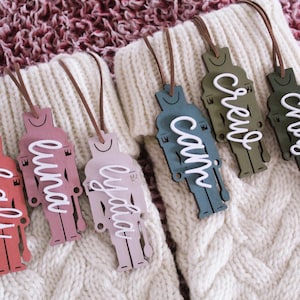 May include: Five wooden nutcracker ornaments with names written in white on each. The ornaments are in various colors: red, pink, light blue, green, and dark green. The ornaments are hanging from white knitted stockings.