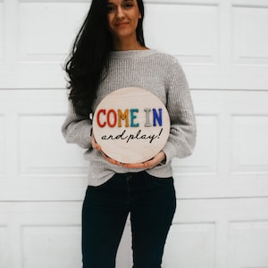 K&ouml;nnte beinhalten: Runder Holzschild mit der Aufschrift "COME IN and play!" in bunten Buchstaben. Das Schild wird von einer Person gehalten, die einen grauen Pullover und dunkle Jeans tr&auml;gt. Der Hintergrund ist ein wei&szlig;es Garagentor.