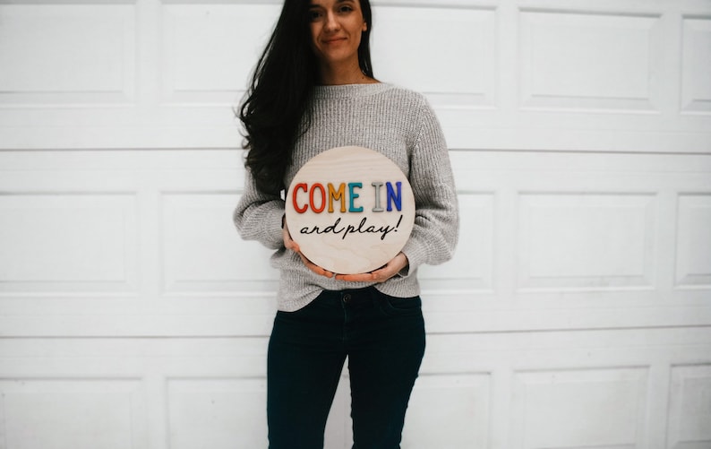 K&ouml;nnte beinhalten: Runder Holzschild mit der Aufschrift "COME IN and play!" in bunten Buchstaben. Das Schild wird von einer Person gehalten, die einen grauen Pullover und dunkle Jeans tr&auml;gt. Der Hintergrund ist ein wei&szlig;es Garagentor.