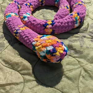 May include: A crocheted snake toy with a pink base and colorful accents. The snake is curled up on a green quilted surface.