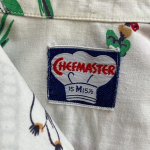 May include: Close-up of a white button-down shirt with a colorful pattern of red, green, and black designs. The shirt has a label that reads "CHEFMASTER" in red letters, with the size "15 M15 1/2" printed below.