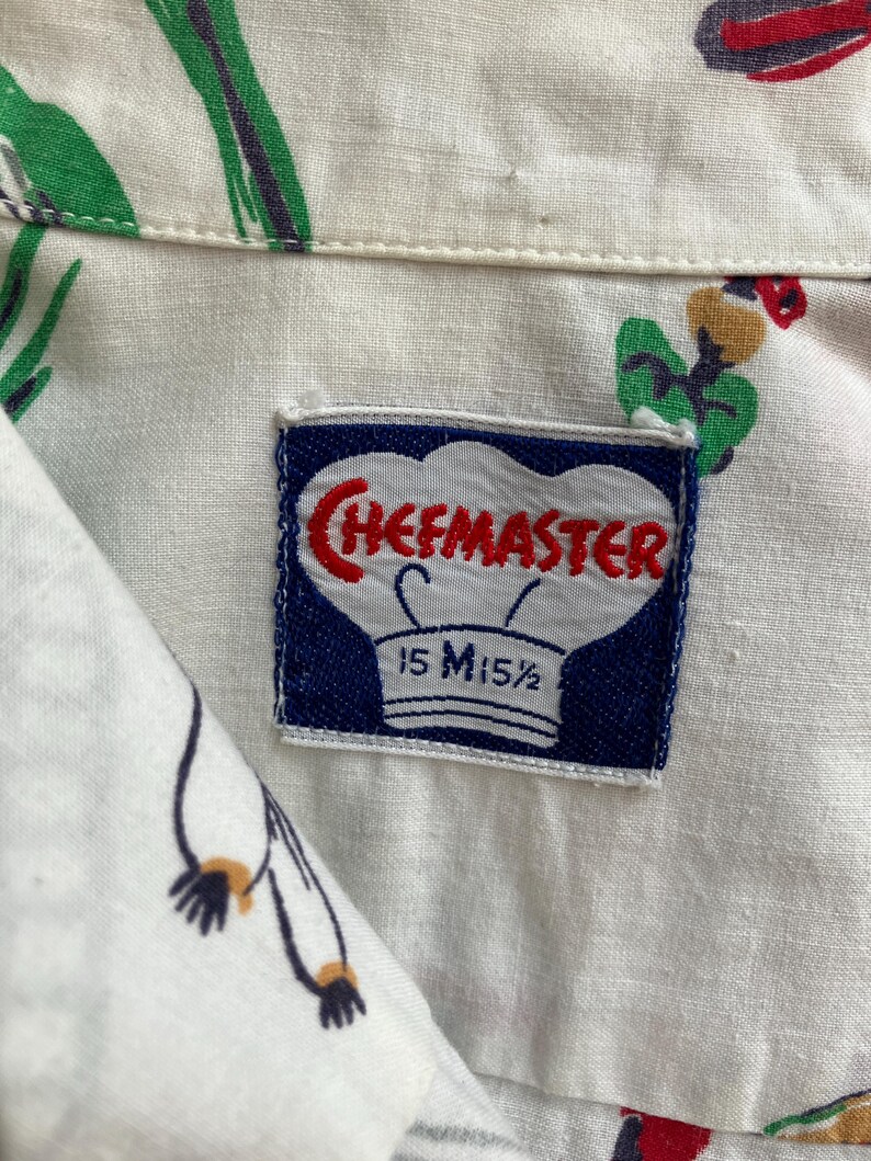 May include: Close-up of a white button-down shirt with a colorful pattern of red, green, and black designs. The shirt has a label that reads "CHEFMASTER" in red letters, with the size "15 M15 1/2" printed below.