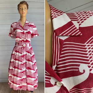 May include: A short-sleeved midi dress featuring a bold red and white graphic pattern of repeating wave-like shapes. The dress has a collared neckline, a full button-down front with white buttons, and a matching fabric belt with a round buckle.