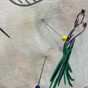 May include: White fabric with a colorful food-themed pattern, featuring illustrations of scallions, and other food items. Three pins with blue and yellow heads are inserted into the fabric.