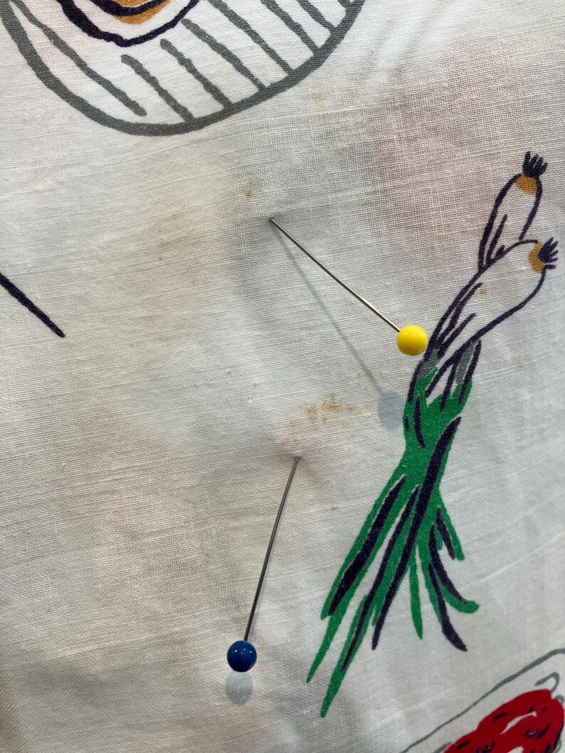 May include: White fabric with a colorful food-themed pattern, featuring illustrations of scallions, and other food items. Three pins with blue and yellow heads are inserted into the fabric.