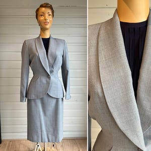 Puede incluir: Un traje gris a medida con falda y chaqueta con un cuello ancho y puntiagudo. La chaqueta tiene un cierre de un solo botón y una silueta ajustada.