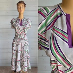 May include: A knee-length dress featuring a light grey base with a repeating pattern of curved, diagonal stripes in shades of green, purple, white, and dark pink. The garment has short sleeves, a round neckline with a solid purple vertical slit detail.