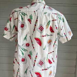 May include: White short-sleeve shirt with a repeating pattern of barbecue-themed illustrations. The design includes red watermelon slices, grilled meats, skewers, and corn on the cob. The shirt is displayed on a mannequin.