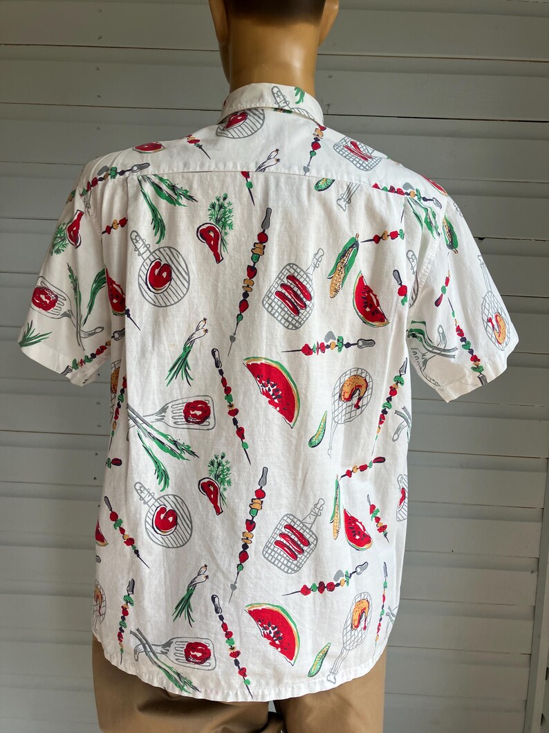 May include: White short-sleeve shirt with a repeating pattern of barbecue-themed illustrations. The design includes red watermelon slices, grilled meats, skewers, and corn on the cob. The shirt is displayed on a mannequin.