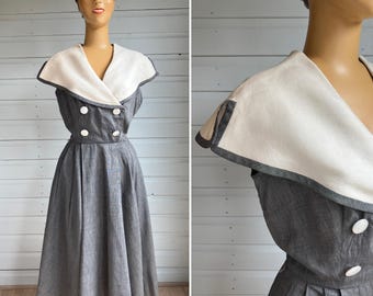 May include: A vintage gray dress with a white collar and gray trim, displayed on a mannequin. The dress features a double-breasted bodice with white buttons and a pleated skirt. The collar is a contrasting white with gray trim.